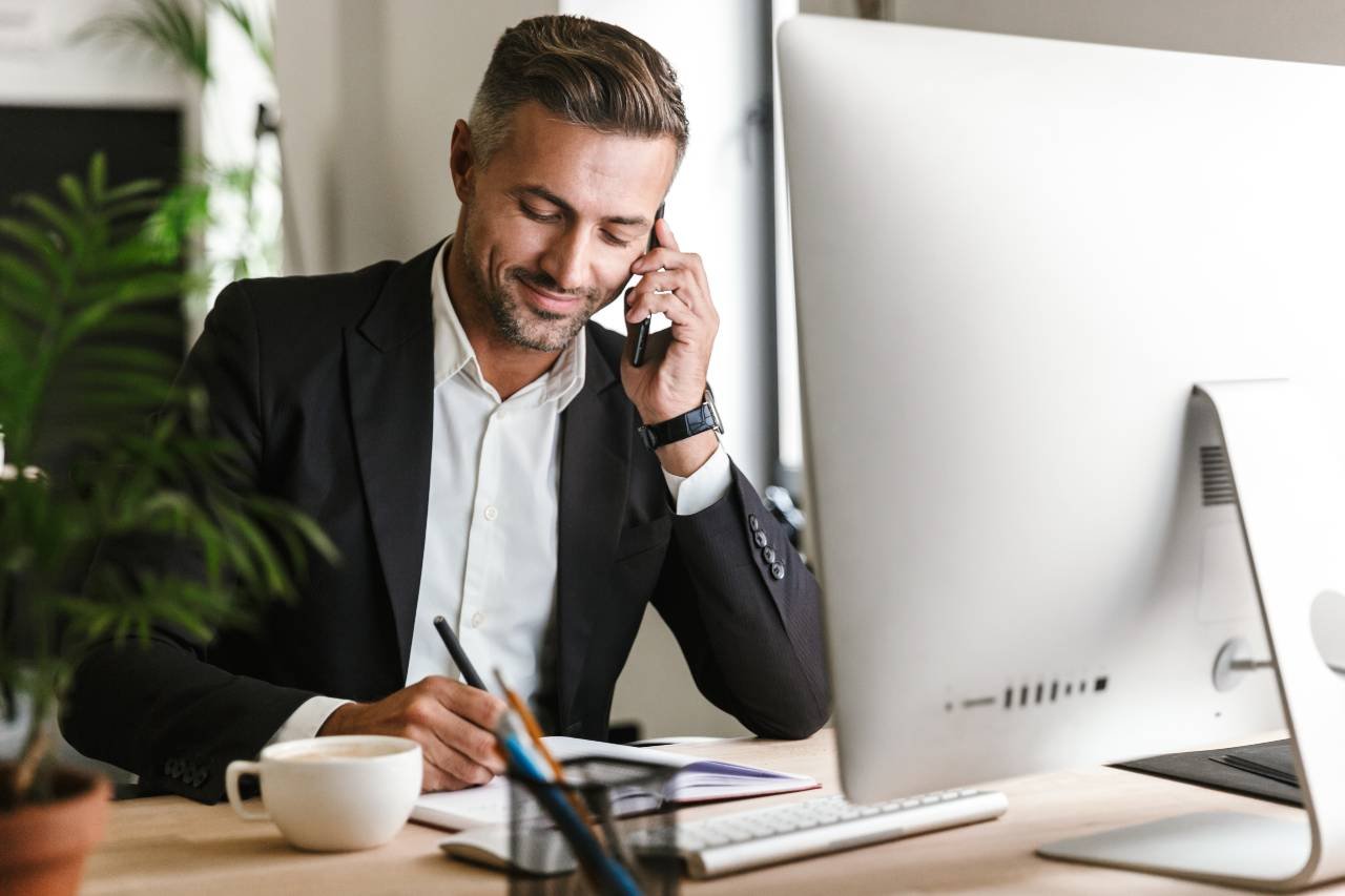 image shows a man in a suit jacket sitting at a desk in front of a computer monitor, taking notes while speaking on the phone. a cup of coffee sits at the desk.