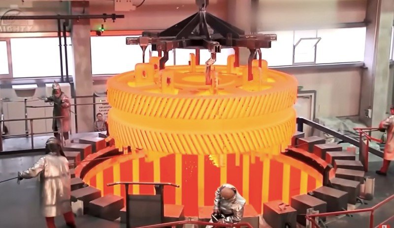 Image shows men working in a factory as a large metal gear is being pulled from a forge and is glowing from heat.